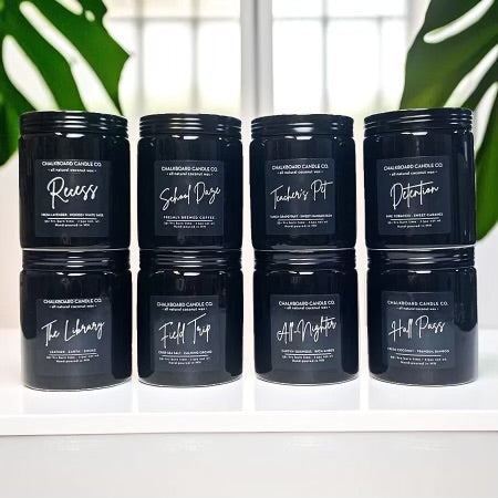 A set of eight black farmhouse candles with white label text stating "Recess, School Daze, Teacher's Pet, Detention, The Library, Field Trip, All-Nighter, and Hall Pass", arranged in two rows on a white surface with a green plant in the background.