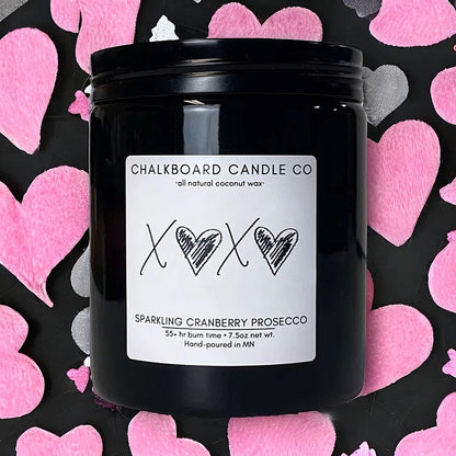 A black jar of Chalkboard Candle Co. sparkling cranberry Prosecco scent candle, with a white label featuring black hearts and the letters 'X' in the center, on a background with pink heart patterns.