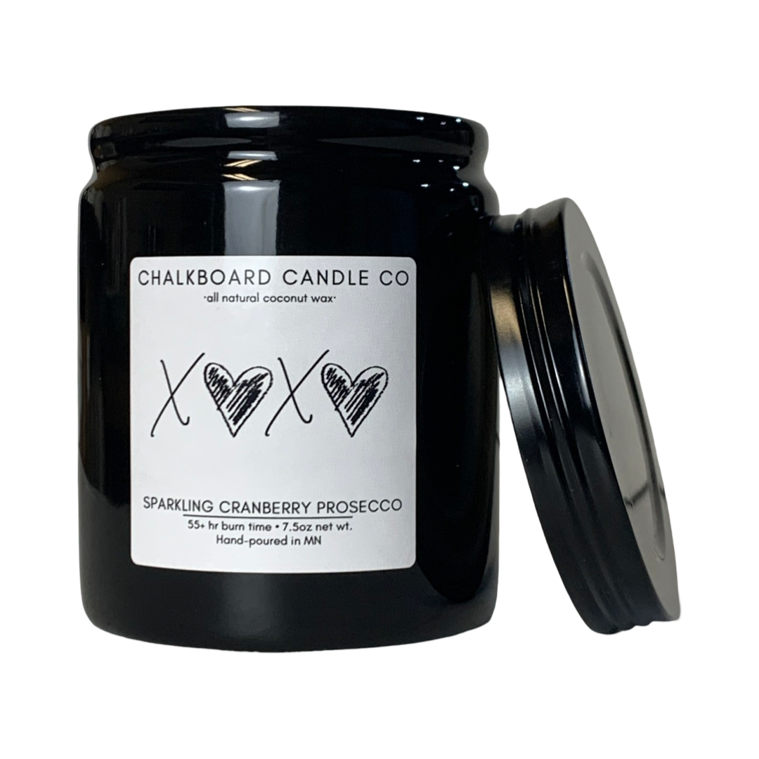 A black jar of Chalkboard Candle Co. sparkling cranberry Prosecco scent candle, with a white label featuring hearts and the letters 'X' in the center on a white background with lid to the side.
