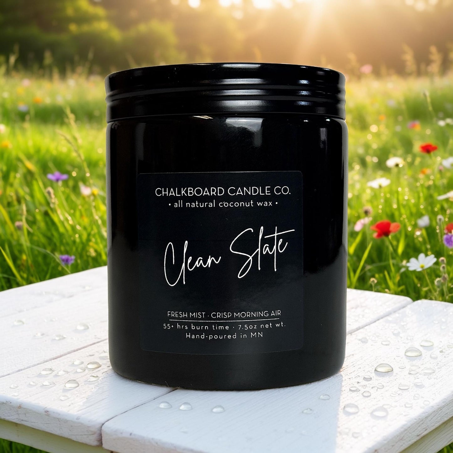 Black Chalkboard Candle Co. jar labeled 'Clean Slate' on dew-speckled white table with blurred wildflower field background