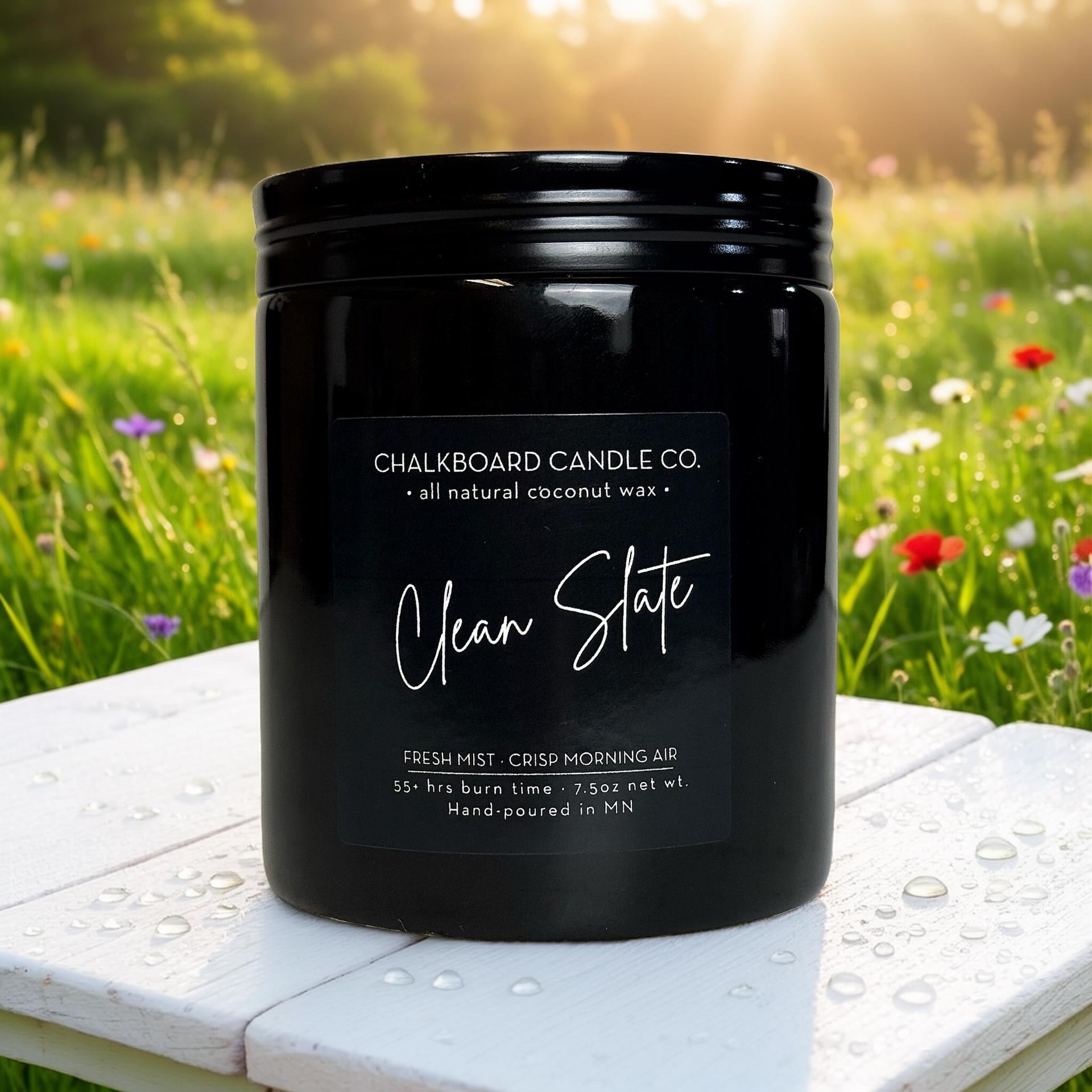 Black Chalkboard Candle Co. jar labeled 'Clean Slate' on dew-speckled white table with blurred wildflower field background
