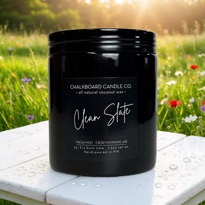 Black Chalkboard Candle Co. jar labeled 'Clean Slate' on dew-speckled white table with blurred wildflower field background