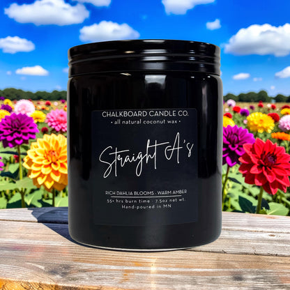 Front view of Straight A’s candle in black ceramic jar showcasing label ‘Straight A’s’ on a wood table with flowers and blue sky
